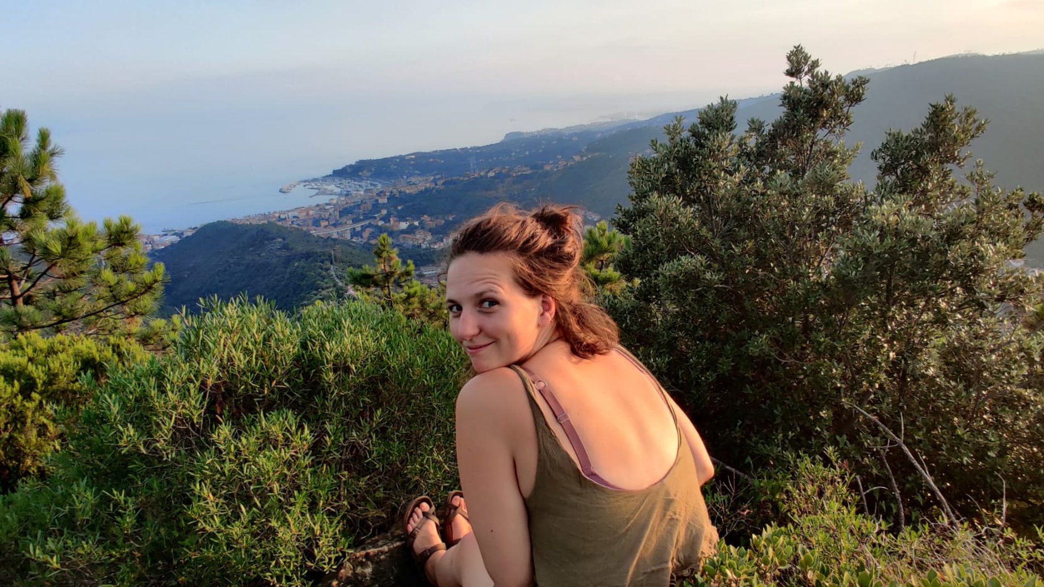 Megan Morgan sitting on a mountain, looking over her shoulder at the camera. In the distance you can see shrubbery and the sea.